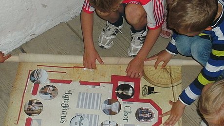 FFM_Schatzsuche_im_Kirchenmuseum Fotoaufnahme der Schatzsuche in der Spitalkirche in Bad Windsheim. Die aufgerollte Schatzkatze ist auf den Holzdielen des Bodens ausgebreitet. Die Kinder sitzen bzw. stehen um die Karte und studieren sie.