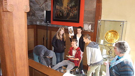 Impressionen aus der Aktion "Und woher kommst du?" Fotoaufnahme der Aktion "Und woher kommst du?" in der Spitalkirche. Im Fokus steht eine Gruppe aus fünf Jugendlichen und einer Erwachsenen. Mittig sitzt ein Junge mit Buch auf einem Stuhl, das Mädchen dahinter hält ein Bild hoch. Im Hintergrund hängt ein Gemälde der Taufe Jesu.
