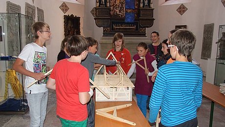FFM_Unter_dem_Dach_der_Kirche“ Fotoaufnahme der Führung „Unter dem Dach der Kirche“ in der Bad Windsheimer Spitalkirche. Eine Gruppe Kinder steht um einen Bierzelttisch. Darauf befindet sich ein Haus-Modell, anhand dessen sie ein Dach konstruieren. Im Hintergrund ist ein Altar zwischen zwei Fenstern der Kirche erkennbar.