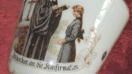 FFM_Vorschau_von_tasse_mit_samt Fotoaufnahme einer Tasse, die auf Samt liegt. Auf der Tasse sind ein Pfarrer und eine Konfirmandin abgebildet. Der Pfarrer im Talar hält eine Bibel in der Hand, aus der er dem Mädchen vorliest. Hinter den beiden ist ein Kreuz erkennbar. Unter dem Bild steht „Andenken an die Konfirmation“